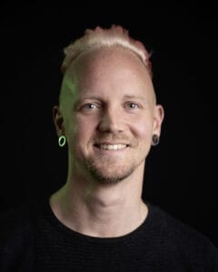 Porträt eines Mannes mit blonden Haaren und Ohrsteckern vor dunklem Hintergrund.