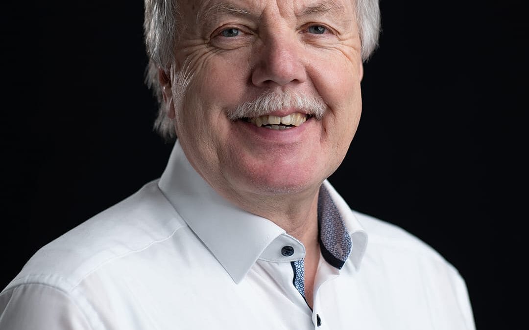 Ein Mann mit grauen Haaren und einem weißen Hemd lächelt in die Kamera vor schwarzem Hintergrund.