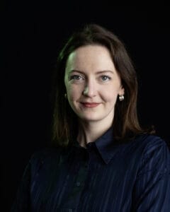 Frau mit langen, braunen Haaren und blauer Bluse, die in die Kamera schaut.