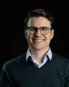 Mann mit Brille und grünem Pullover, der in die Kamera lächelt.