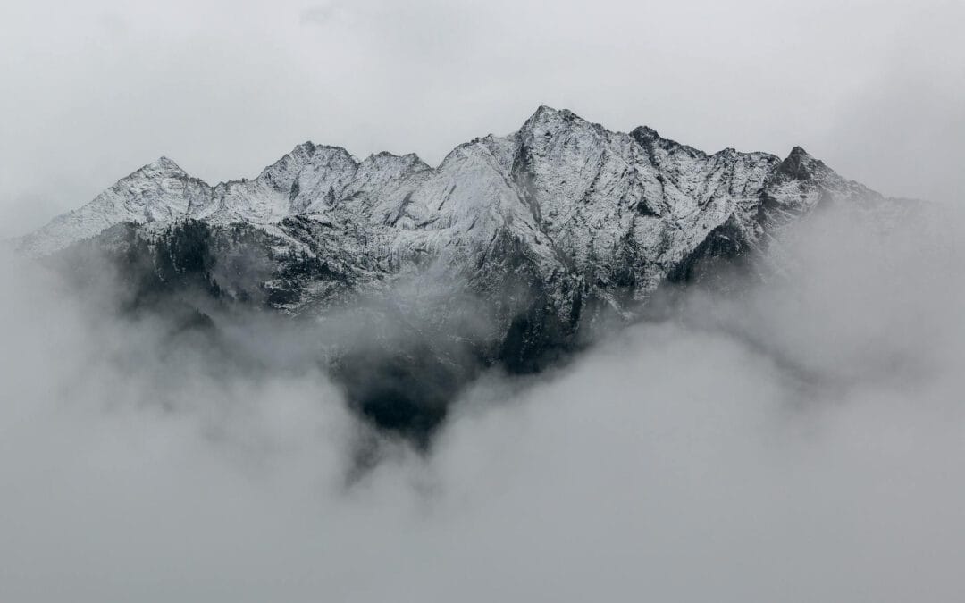 Schneebedeckte Bergspitzen, die teilweise von Wolken umgeben sind, mit grauem Himmel im Hintergrund.