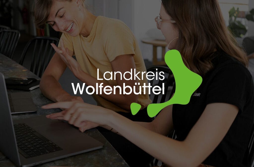 Zwei Frauen sitzen an einem Tisch und interagieren während sie auf einen Laptop schauen, mit Logo Landkreis Wolfenbüttel im Vordergrund.