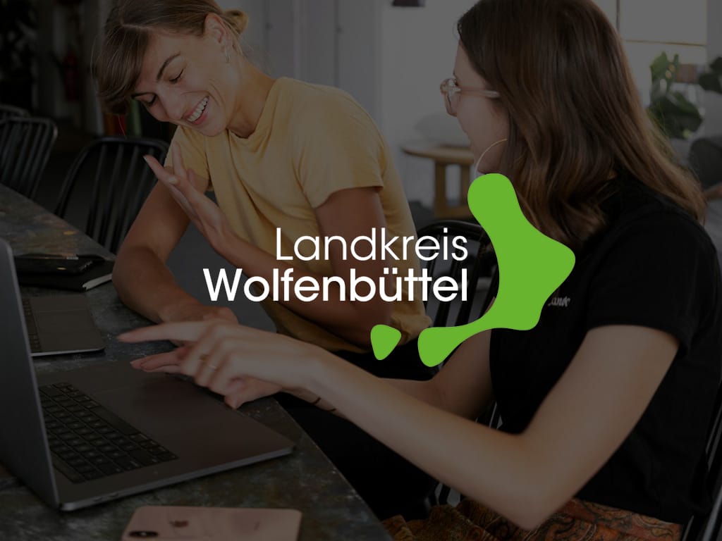 Zwei Frauen sitzen an einem Tisch und interagieren während sie auf einen Laptop schauen, mit Logo Landkreis Wolfenbüttel im Vordergrund.