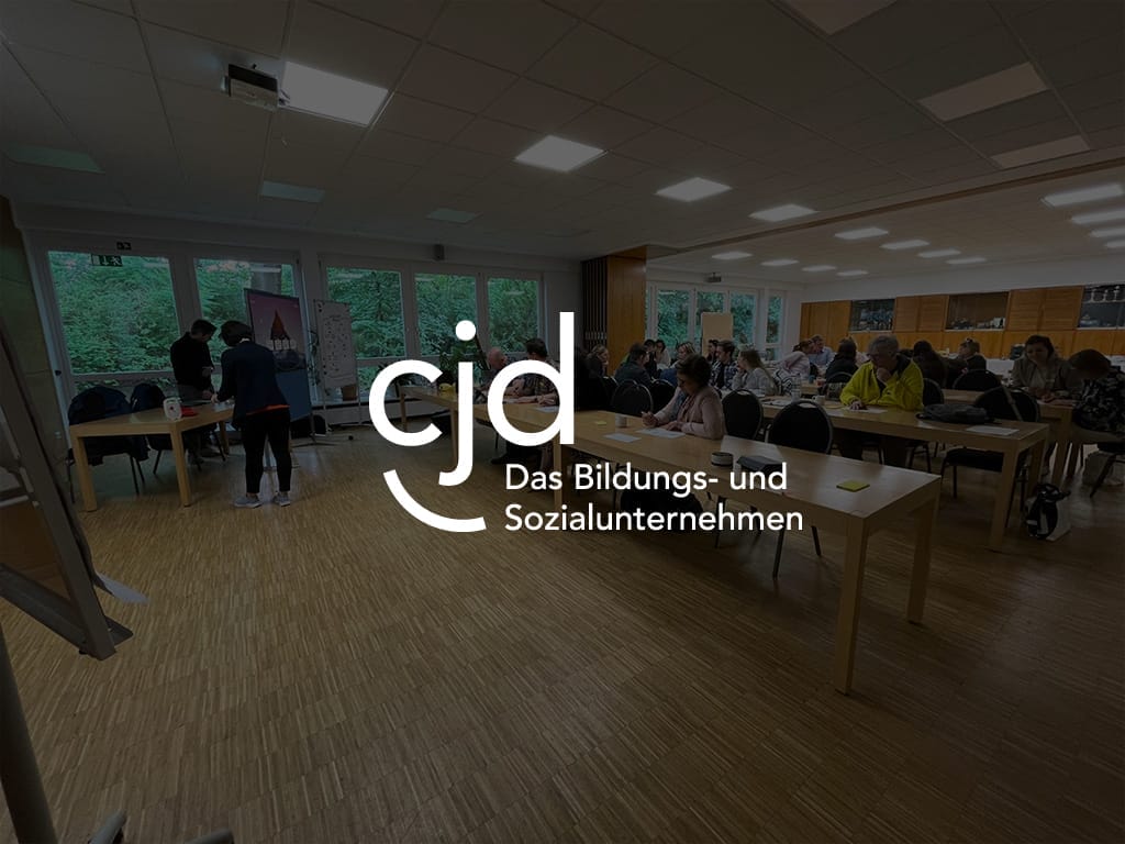 Teilnehmer bei einer Veranstaltung des CJD Bildungs- und Sozialunternehmens in einem Raum mit Tischen und Stühlen.