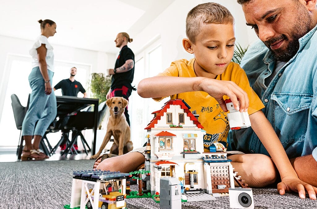 Ein Vater und sein Sohn spielen mit einem Lego-Haus auf dem Boden in einem modernen Raum.