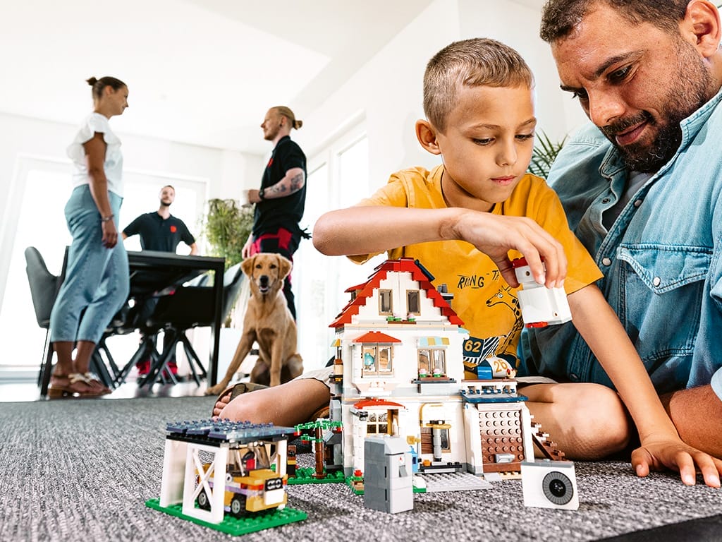 Ein Vater und sein Sohn spielen mit einem Lego-Haus auf dem Boden in einem modernen Raum.