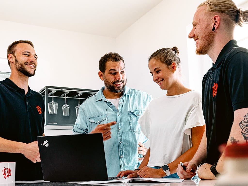 Vier Personen diskutieren an einem Tisch mit Laptops und Notizen in einem hellen Raum.