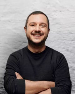 Lächelnder Mann mit kurzen Haaren und schwarzem Pullover, der seine Arme verschränkt.