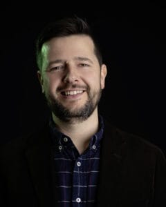 Porträt eines Mannes mit Bart, der in einem dunklen Anzug und karierten Hemd lächelt.