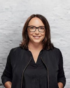 Frau mit Brille und braunen Haaren, die in schwarzer Kleidung vor einer hellen Wand steht.