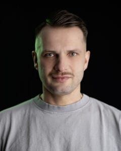 Porträt eines Mannes mit kurzem Haar und einem grauen T-Shirt vor schwarzem Hintergrund.