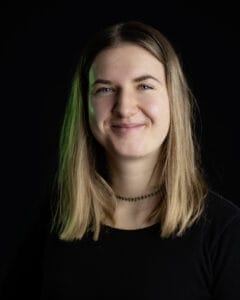 Porträt einer Person mit langen, glatten Haaren und einem freundlichen Lächeln vor schwarzem Hintergrund.