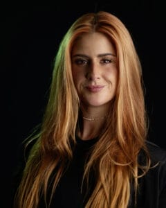 Porträt einer Frau mit langen, roten Haaren, die in die Kamera lächelt.