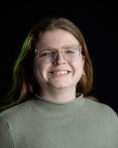 Porträt einer Person mit langen Haaren und Brille, die in die Kamera lächelt.