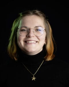 Lächelnde Frau mit Brille und einem schwarzen Rollkragenpullover, die eine Kette mit einem Anhänger trägt.