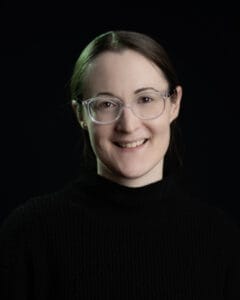 Porträt einer Person mit Brille, die einen schwarzen Pullover trägt und in die Kamera lächelt.