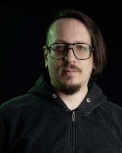 Porträt eines Mannes mit Brille und Kapuzenpullover vor dunklem Hintergrund.