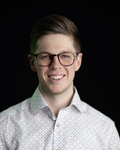 Mann mit Brille und einem weißen Hemd mit Punkten vor schwarzem Hintergrund.