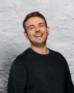 Lächelnder Mann in schwarzem Pullover vor einer grauen Wand.