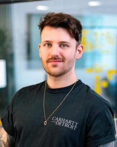 Mann mit kurzem Haar und Bart trägt ein schwarzes Carhartt T-Shirt und eine goldene Kette.