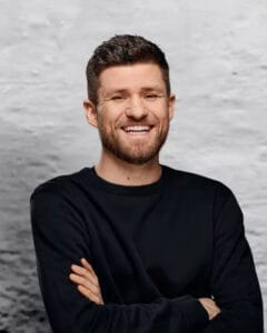 Lächelnder Mann mit kurzen Haaren und schwarzem Pullover, der mit verschränkten Armen posiert.