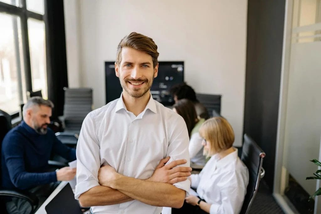 Mann mit braunen Haaren und weißem Hemd steht in einem Büro mit lächelndem Gesicht.